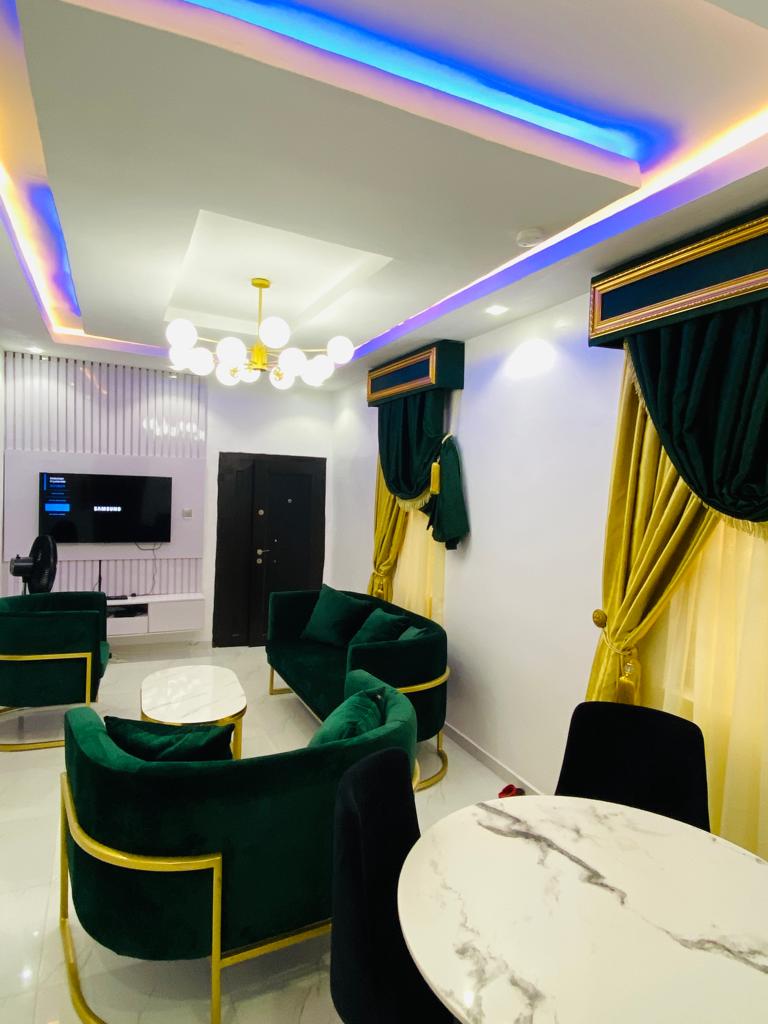 The living area is adorned with modern furnishings, featuring a plush green sofa set and stylish chairs. Elegant curtains frame the windows, while a marble-topped table enhances the decor. Colorful LED lighting adds an inviting ambiance, complemented by a wall-mounted TV.
