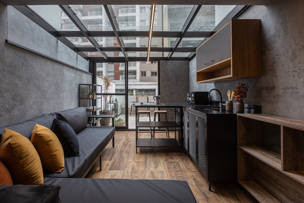 A modern studio interior features a cozy seating area with dark cushions placed on a streamlined couch. A kitchen counter with sleek cabinetry is visible, alongside a small dining table and chairs. Large windows allow natural light to fill the space, offering views of the outside.