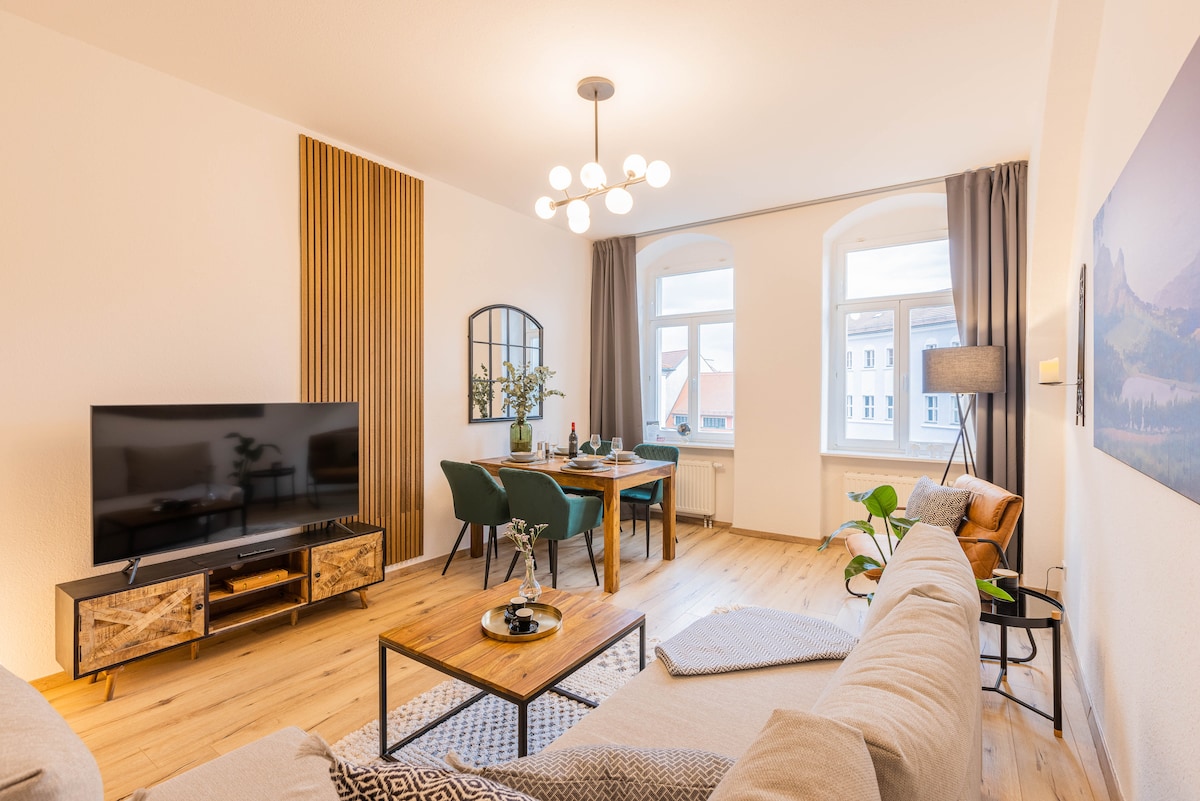A bright living area features a spacious sofa and a wooden coffee table. A large TV is set on a stylish media unit. The dining area includes a wooden table with seating for four. Sunlight streams in through the two arched windows, creating a welcoming atmosphere.