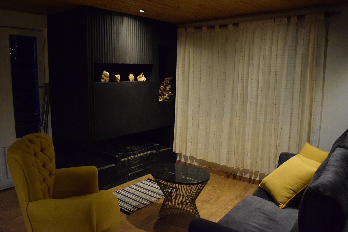 A cozy living area features a dark fireplace with decorative elements above. Two comfortable chairs, one yellow and one gray, are positioned around a glass-top coffee table. Soft curtains filter the evening light, enhancing the intimate ambiance of the space.