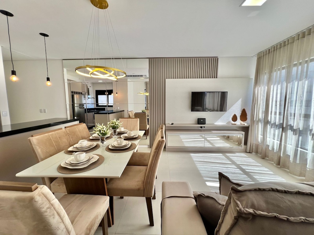 An elegant dining area features a light-colored table set for six, accompanied by plush chairs. A modern entertainment unit with a television is positioned against a wall, while soft sunlight filters through sheer curtains, highlighting the spaciousness of the room.