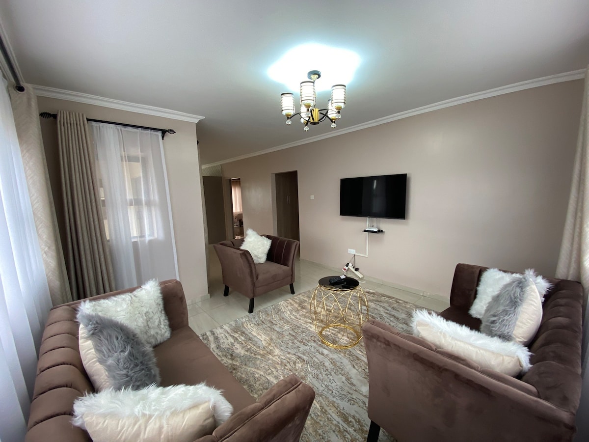 A modern living area is furnished with two plush armchairs and a soft area rug. A glass coffee table sits at the center, while a wall-mounted television provides entertainment. Natural light filters through sheer curtains, adding warmth to the neutral-toned space.