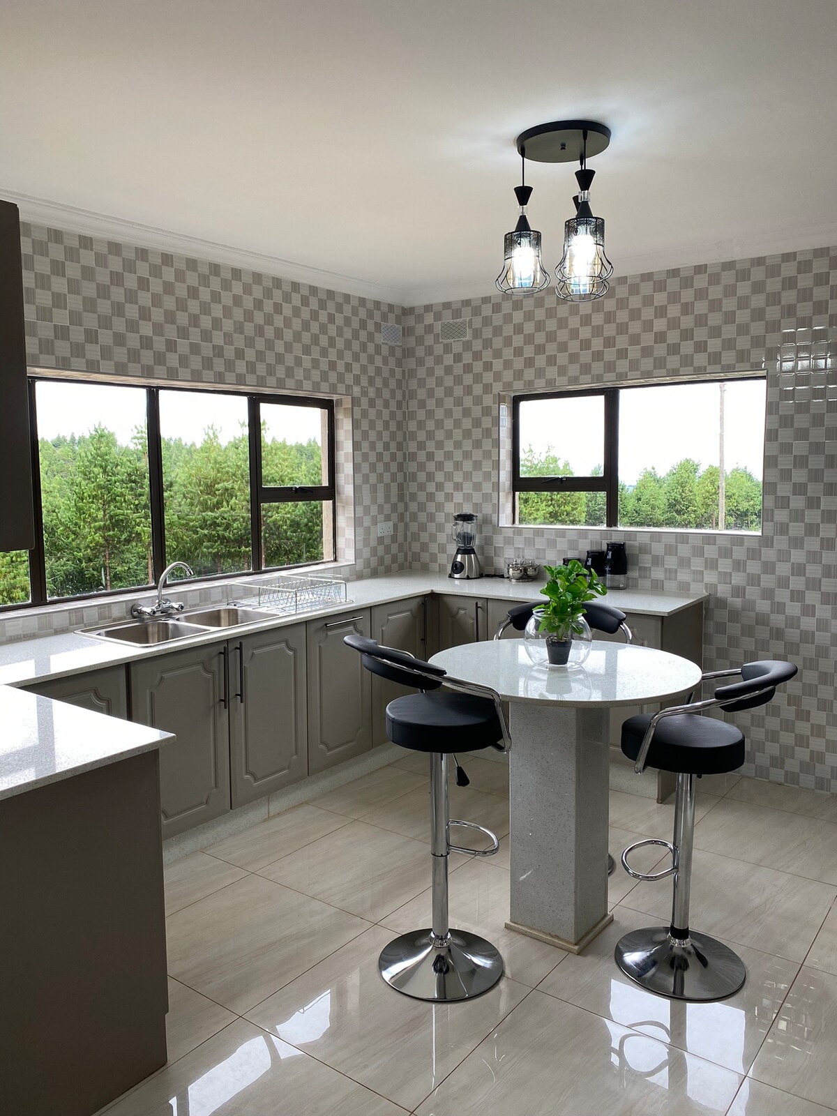 A modern kitchen is depicted, featuring light-colored cabinetry and a checkered wall pattern. A round dining table with four black bar stools is positioned near large windows, offering views of the surrounding greenery. Stainless steel appliances and a sleek sink are also present.