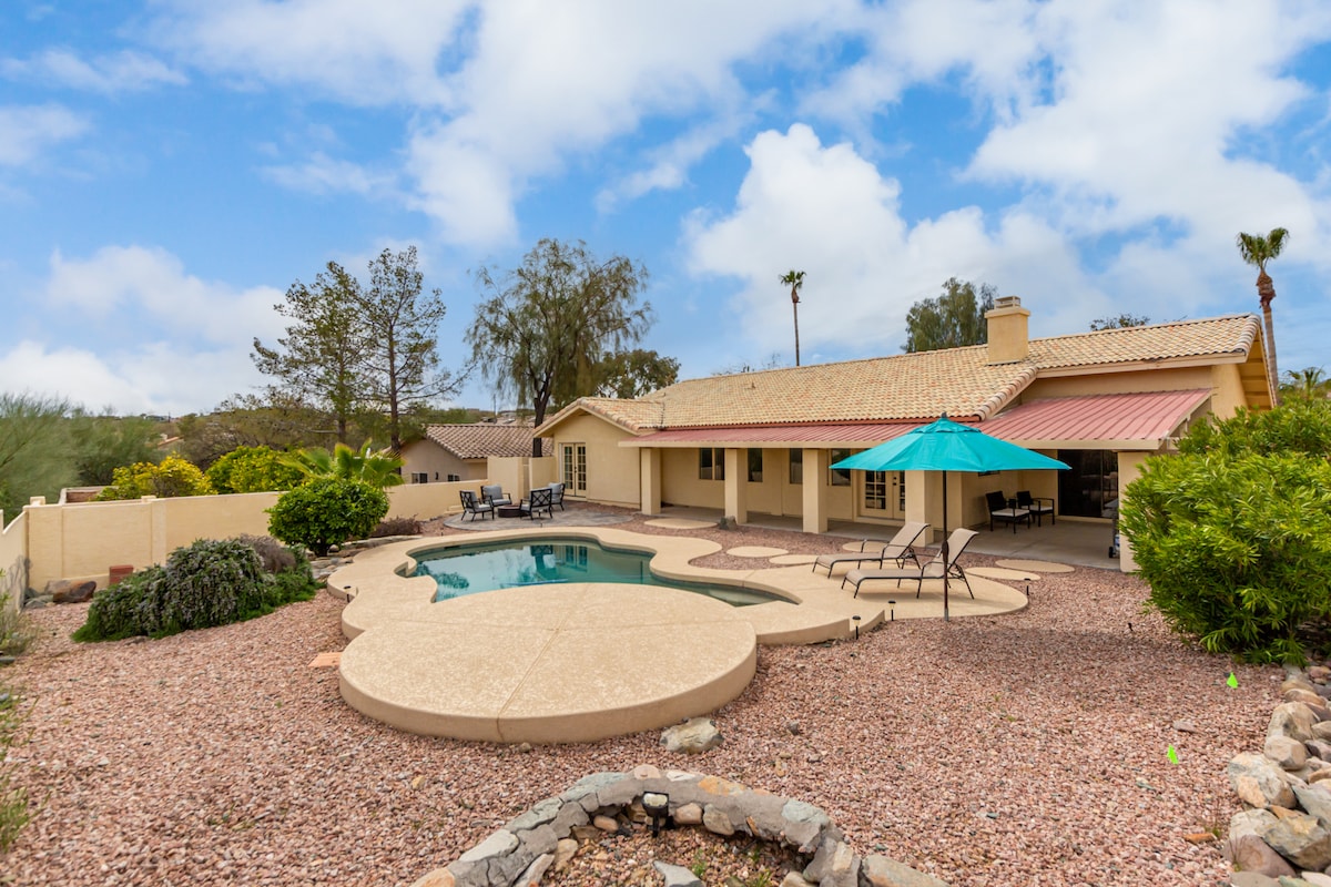 The exterior of the home is presented, featuring a spacious backyard with a swimming pool shaped like a natural oasis. A covered patio area with a blue umbrella is visible, alongside a seating arrangement. Surrounding desert landscaping complements the tranquil setting, with low-maintenance rock beds and a mix of shrubs.