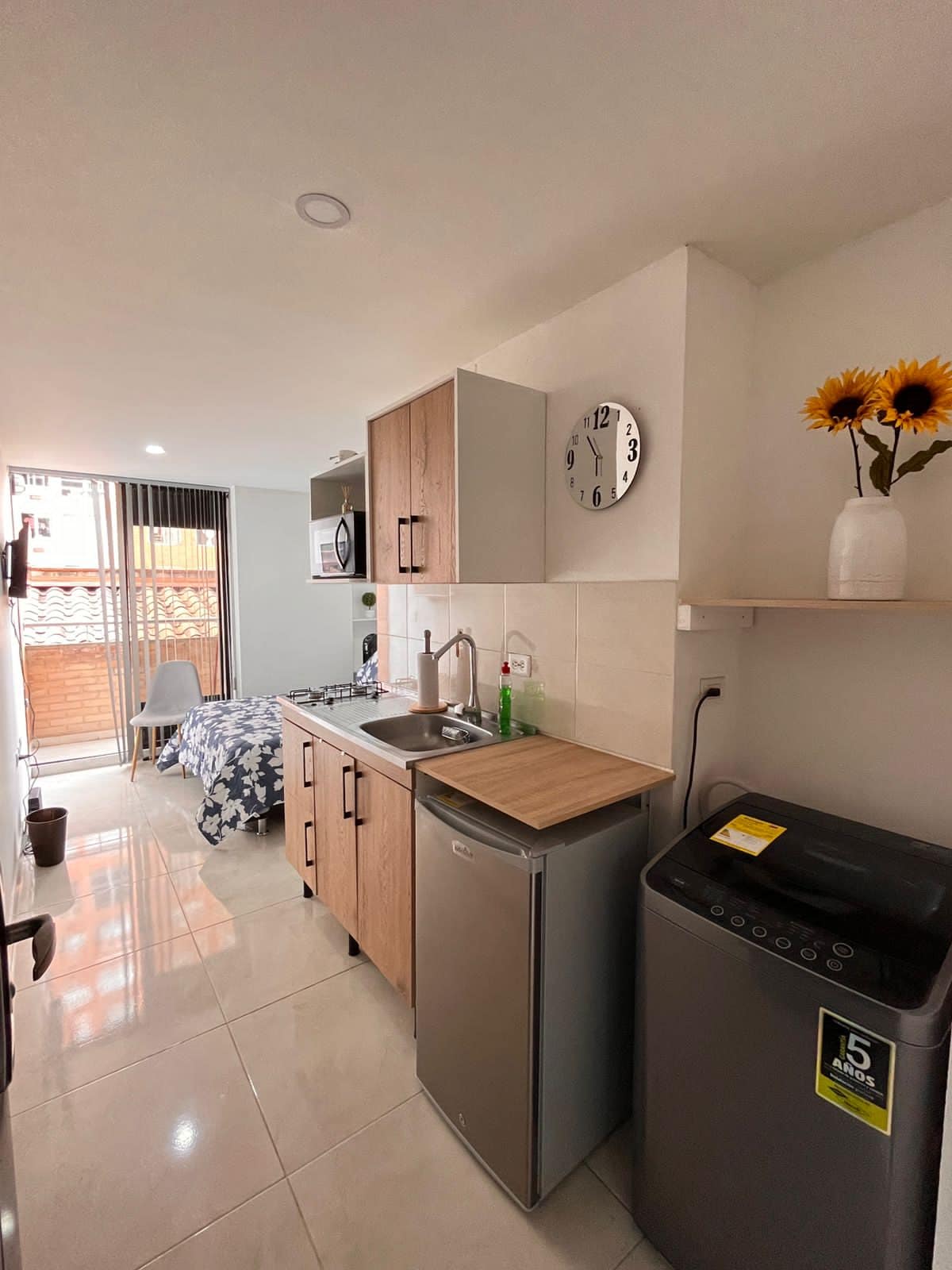 A compact kitchen area showcases modern cabinetry and essential appliances, including a sink, microwave, and refrigerator. A small dining space is positioned beside a window, with natural light illuminating the space. Decorative elements include a vase of sunflowers on a shelf, adding a touch of brightness.