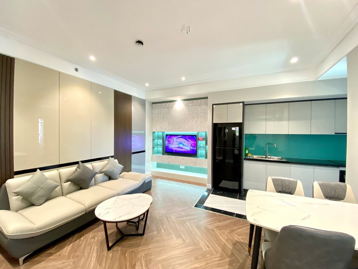 A modern living space is shown, featuring a light-colored sectional sofa with decorative pillows. A sleek, white coffee table sits in front. The kitchen area includes a black refrigerator and a turquoise backsplash. A flat-screen TV is mounted on the wall, enhancing the room's functionality.