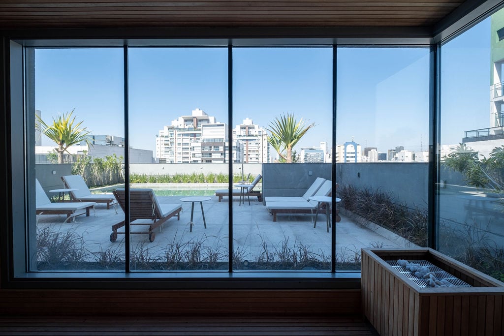 Large glass windows provide a view of a spacious outdoor area, featuring lounge chairs and sun loungers by a swimming pool. Lush greenery surrounds the pool area, complementing the urban backdrop of São Paulo's skyline in the distance.