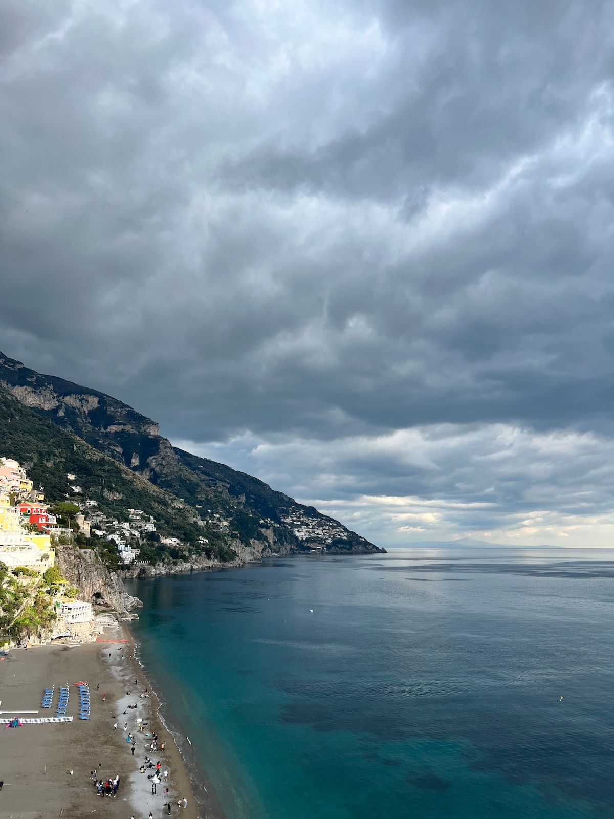 A coastal view of the Positano shoreline is presented, with gentle waves lapping against the sandy beach. Rolling hills rise steeply on one side, adorned with colorful buildings. A cloudy sky casts shadows over the scene, creating a serene atmosphere over the water.