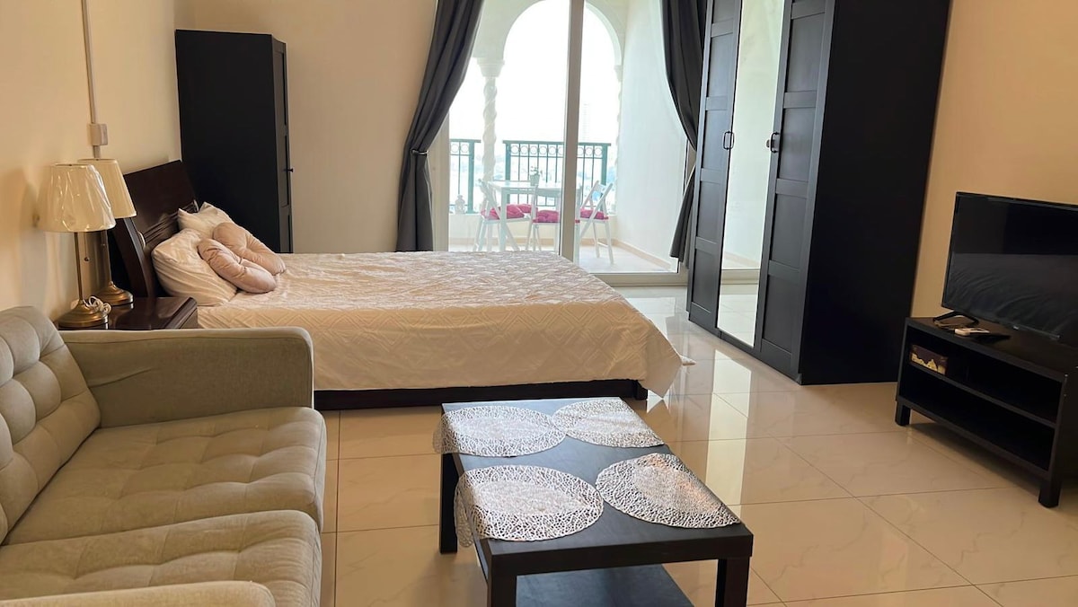 A cozy studio flat features a spacious bed with a light quilt, accompanied by two decorative pillows. A grey sofa is positioned nearby, alongside a dark wood coffee table adorned with circular coasters. Floor-to-ceiling windows provide access to a balcony with a small outdoor seating arrangement.