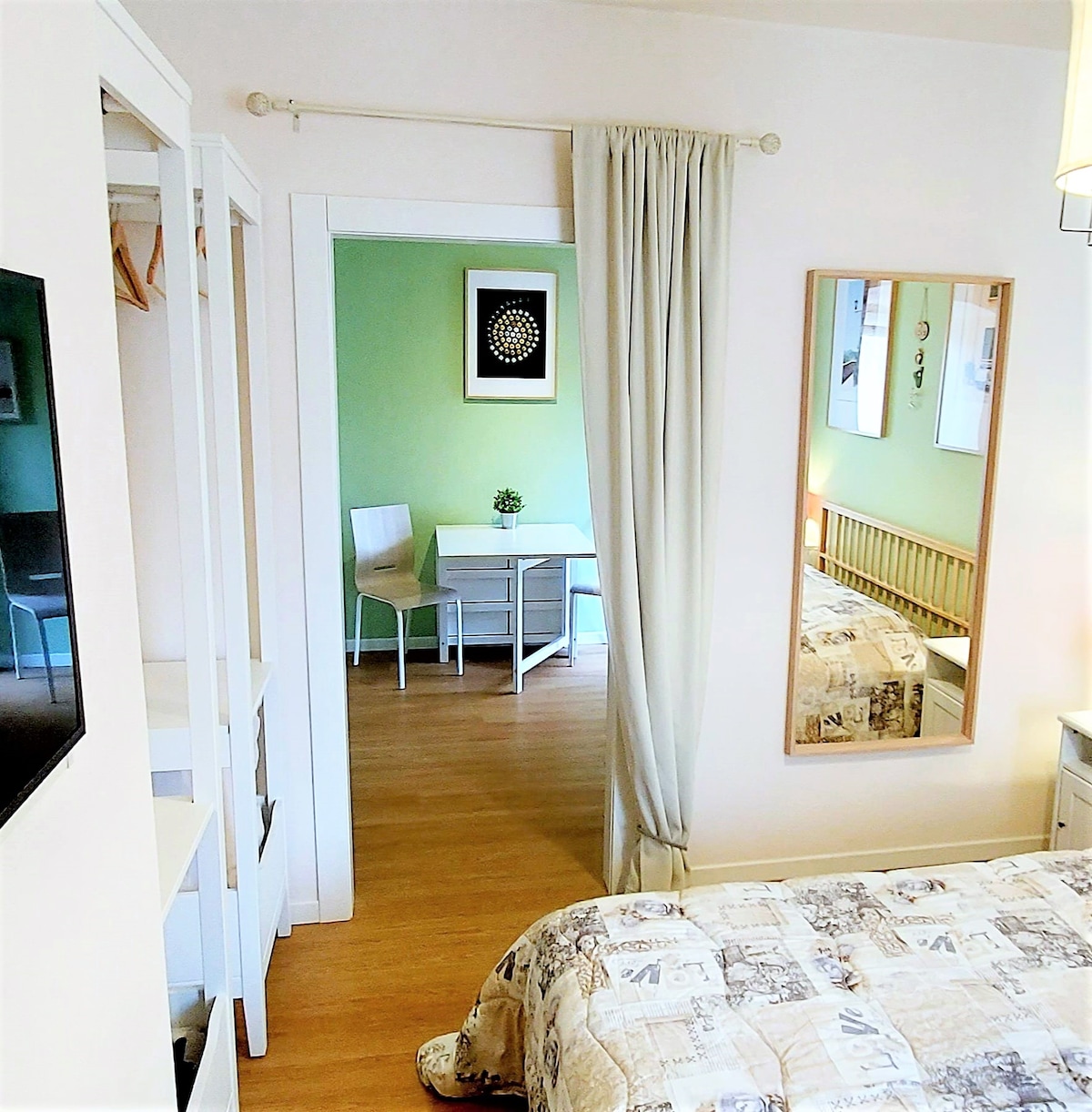 A cozy bedroom is seen with a large bed dressed in a patterned coverlet. A full-length mirror reflects a small dining area in the background, featuring a table and two chairs against a green wall. Natural light enters through the adjacent room.
