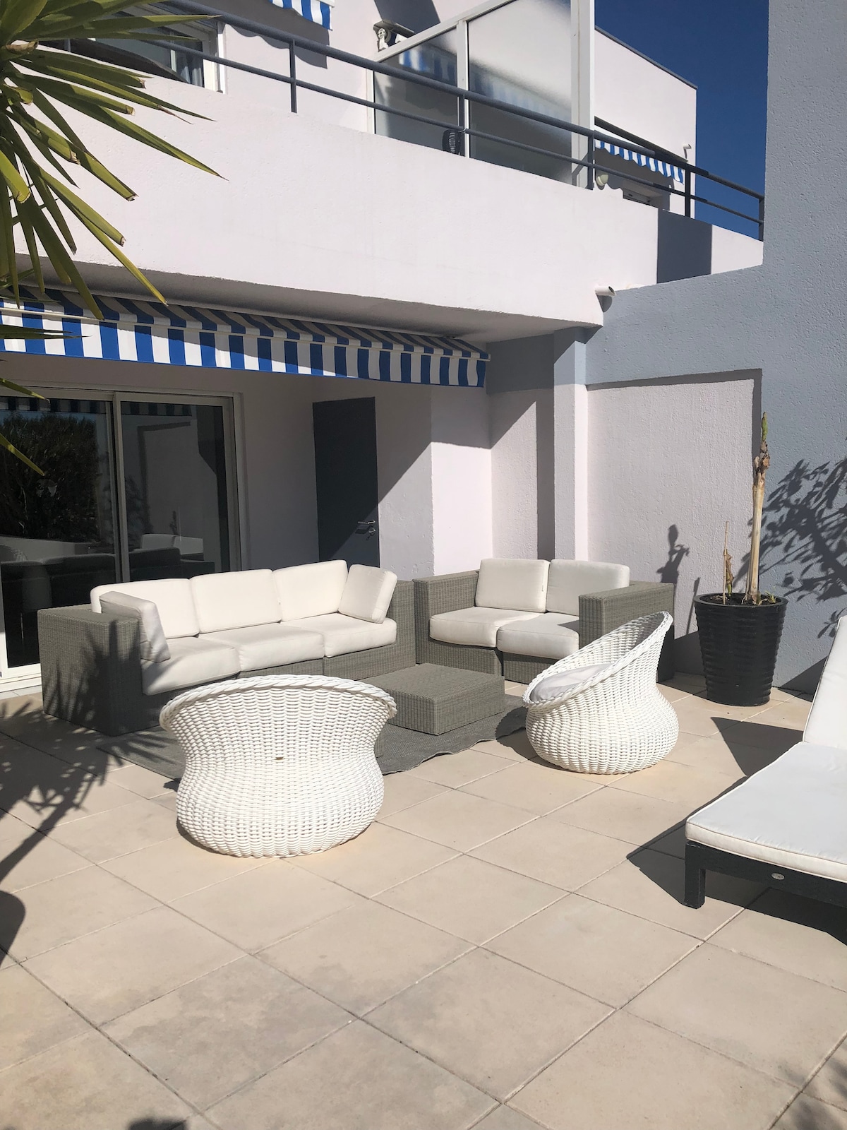 A spacious terrace is furnished with a light-colored sofa set and two woven chairs, surrounded by neutral tiled flooring. Potted plants add a touch of greenery, while blue and white striped awnings provide shade. Glass doors lead into the interior of the apartment.