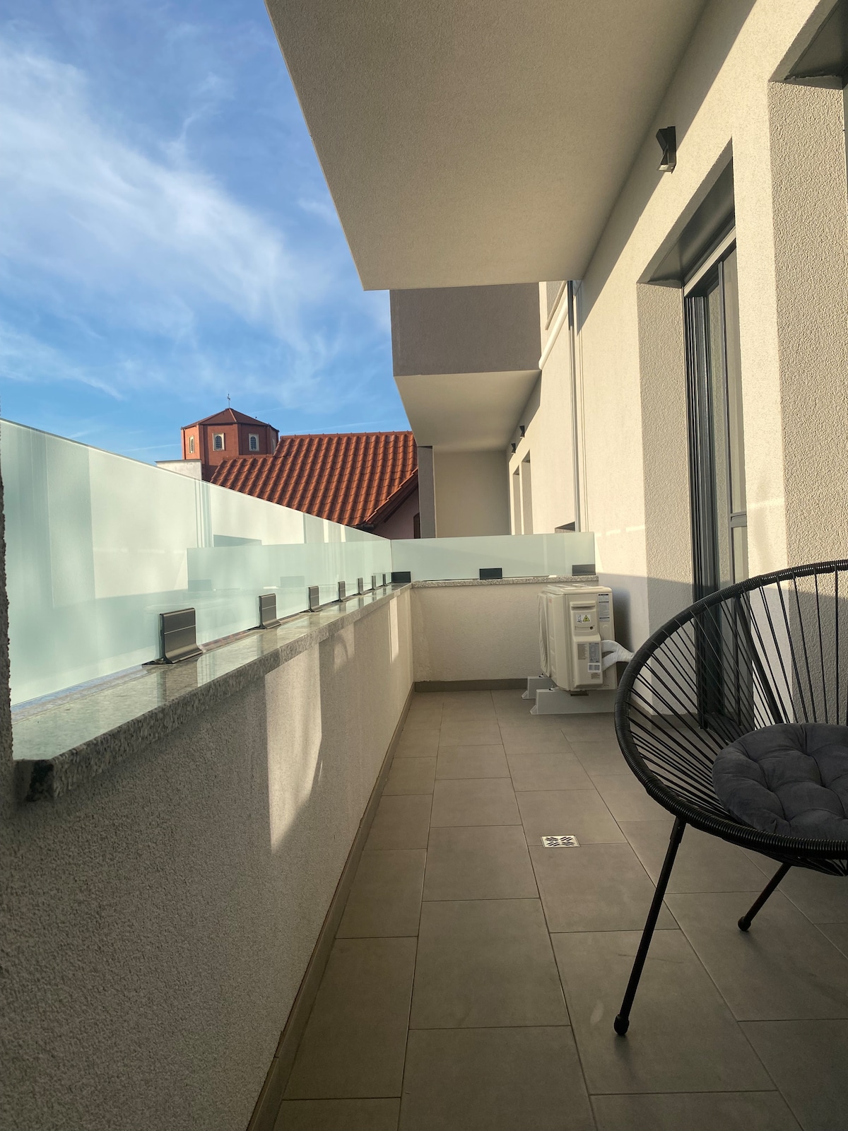 A private balcony is showcased, featuring a sleek glass railing. Sunlight casts shadows on the tiled floor, with an accent chair positioned for relaxation. The surrounding architecture, visible in the background, adds a touch of character to the space.