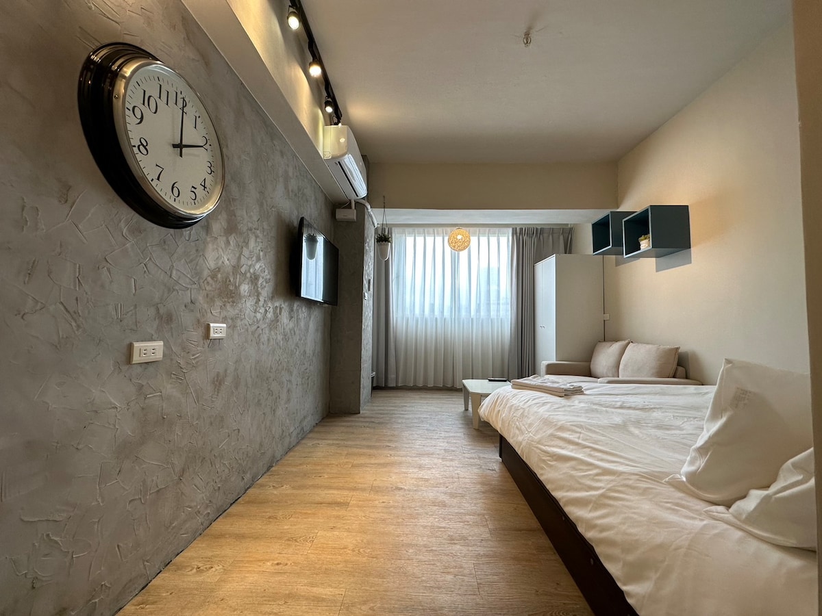 A spacious room features a large double bed, a minimalistic white sofa, and a television mounted on the wall. Natural light enters through sheer curtains, complementing the neutral color palette of the textured wall and wooden flooring. A stylish clock adds a functional accent.