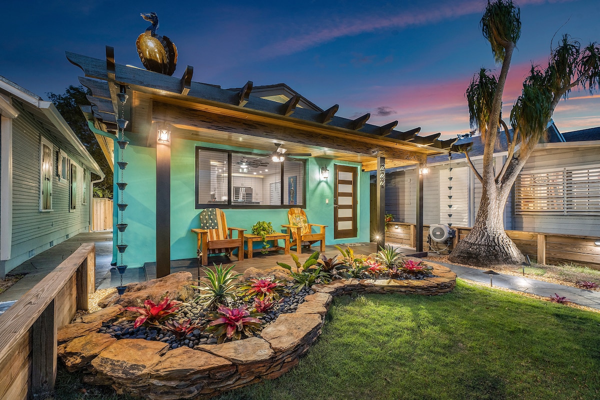 A charming exterior features a vibrant turquoise facade, complemented by a cozy seating area with two chairs on the front porch. Lush landscaping with colorful plants surrounds the stone pathway leading to the entrance, while decorative lighting adds a welcoming touch as dusk settles.