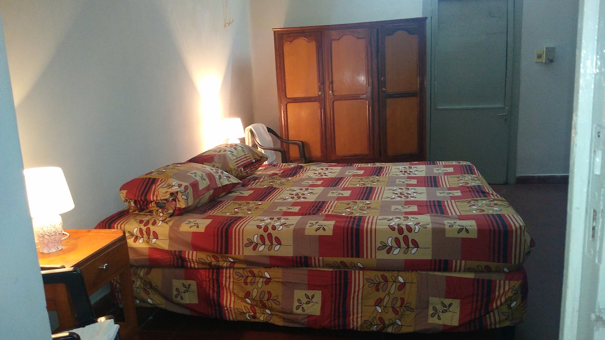A spacious sleeping area is presented with a large bed featuring a patterned duvet. A wooden wardrobe is positioned against the wall, and a small bedside table holds a lamp. A chair is visible near the bed, providing additional seating.