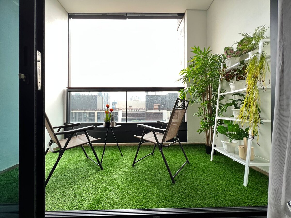 A small balcony features two folding chairs and a vibrant green carpet that mimics grass. A white shelving unit holds various plants, while a flower pot adds a touch of color. The view outside includes distant buildings, enhancing the urban ambiance.