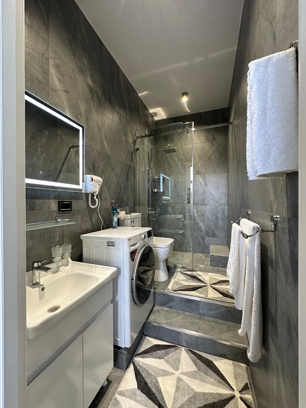 A modern bathroom features sleek gray tiles and a walk-in shower with glass doors. A white vanity with a sink and a washing machine are included. Two towels hang neatly on a rack, while a geometric-patterned rug adds subtle contrast to the floor.
