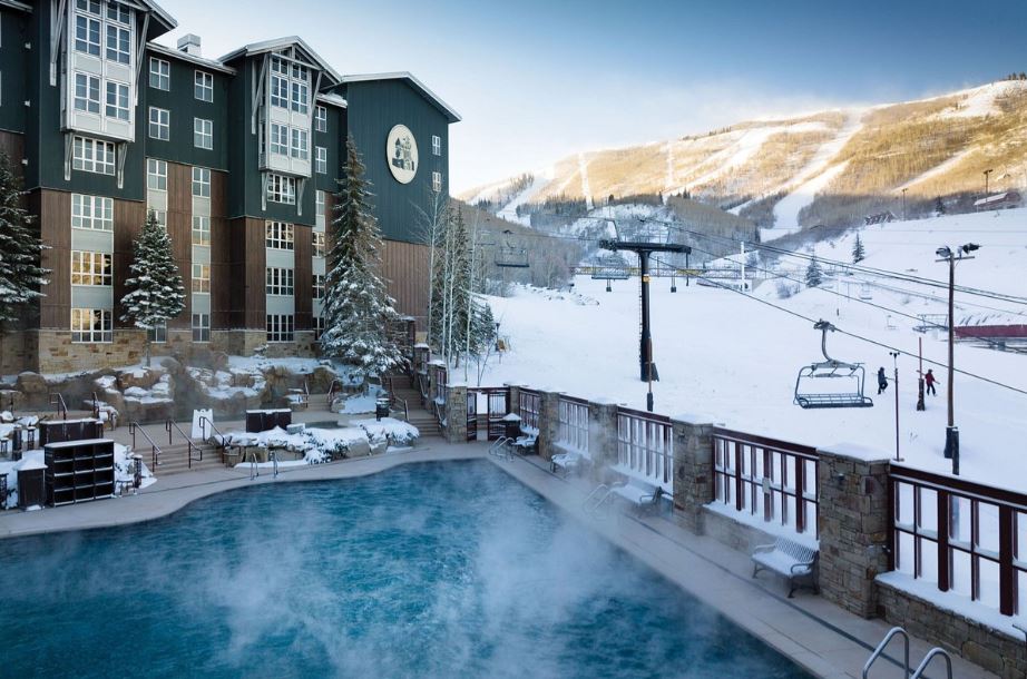 An outdoor heated pool is visible, surrounded by a stone deck and glass railing. Snow-covered mountains and a ski lift are seen in the background, with the resort building featuring a mix of wood and green facade reflected in the serene water.