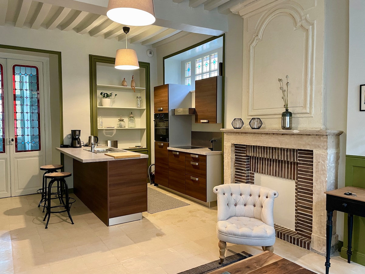 The kitchen and dining area features a modern design with wooden cabinetry and a contrasting light countertop. A small dining table with stools is positioned near the kitchen. The room is illuminated by soft pendant lighting, while a decorative fireplace adds an elegant touch.