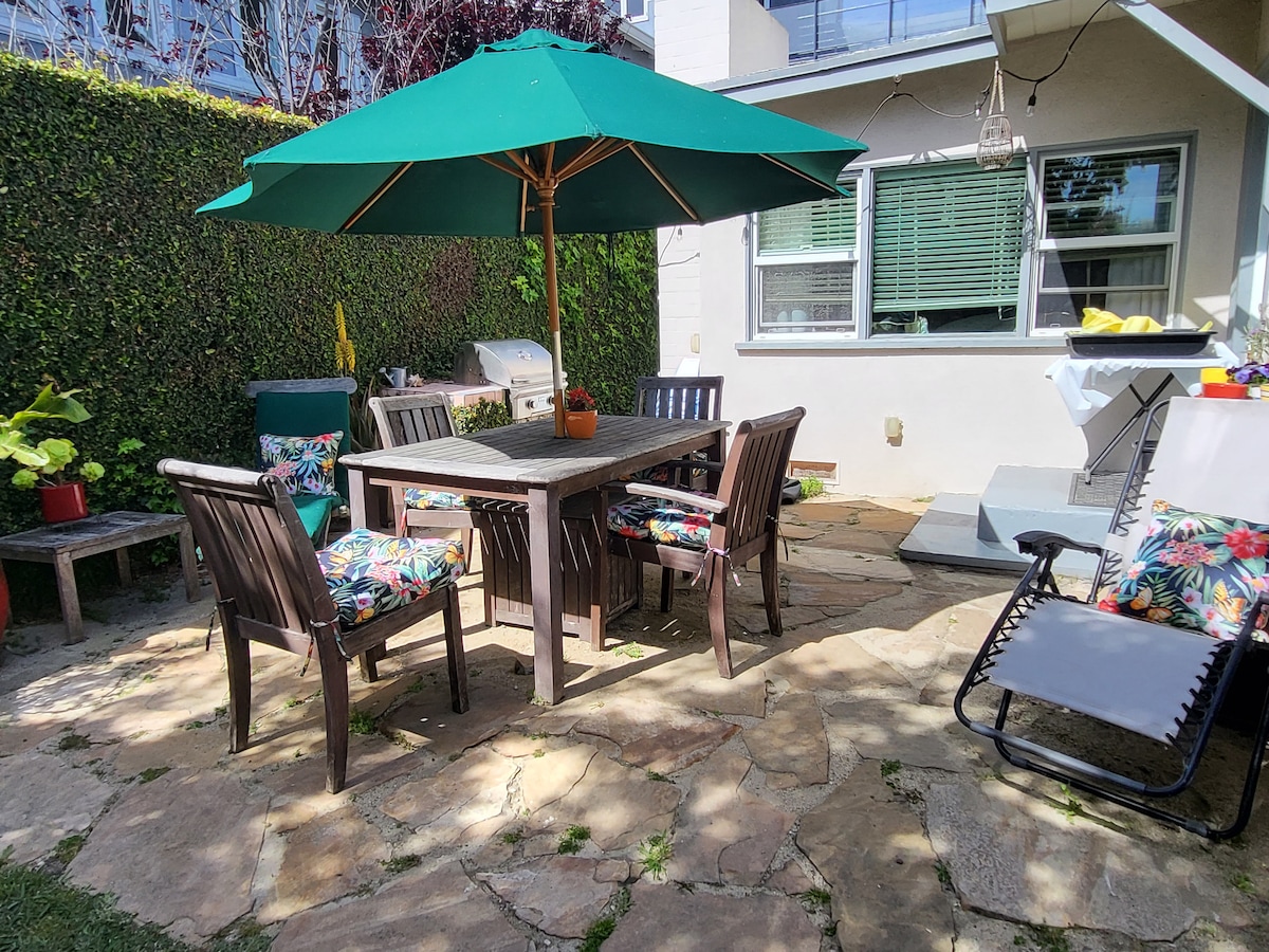 An outdoor patio area features a wooden table surrounded by six chairs, each adorned with colorful cushions. A large green umbrella provides shade, and a gas grill is positioned nearby. Lush greenery lines the perimeter, adding privacy and a sense of tranquility.