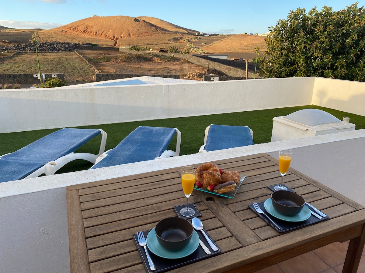 A patio table is set for breakfast with two plates, glasses, and silverware, overlooking a scenic view of rolling hills. Sun loungers are positioned nearby, enhancing the outdoor space. The area is bathed in natural light, with a backdrop of greenery and distant volcanic landscapes.