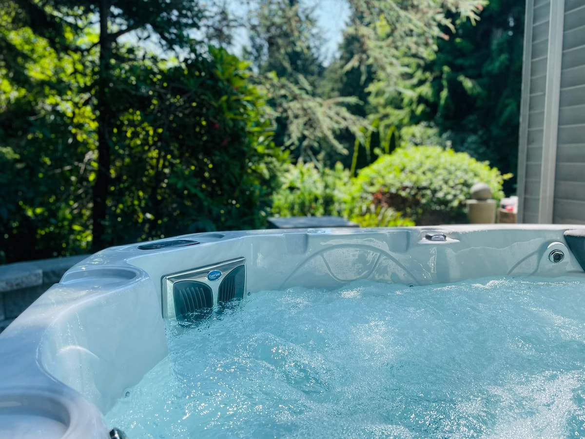 A hot tub is positioned outdoors, surrounded by lush greenery and trees. Sunlight reflects off the water, creating a relaxing ambiance. The hot tub's controls are visible, enhancing the inviting outdoor space.