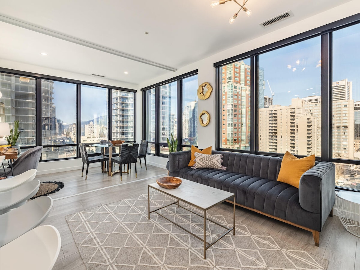 An open-concept living space features large windows that provide expansive views of the downtown skyline. Modern furnishings include a black velvet couch with orange accents and a coffee table centered on a textured area rug. A dining area is visible, complementing the overall design.