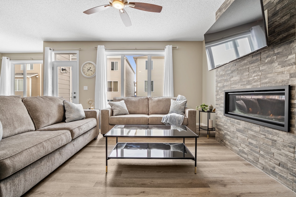 A spacious living area features two comfortable sofas facing a modern fireplace surrounded by stone. A glass coffee table sits in the center, reflecting natural light from large windows adorned with sheer curtains. Light wood flooring adds warmth to the space.