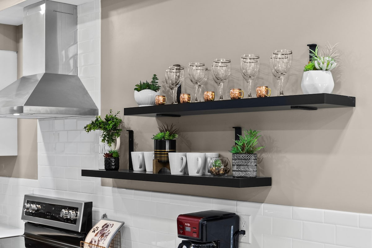 Open shelving displays a variety of glassware and decorative plants. The top shelf features clear glasses, while the lower shelf holds white mugs alongside small potted foliage and decorative containers. A modern kitchen is seen beneath the shelves, enhancing functionality and style.
