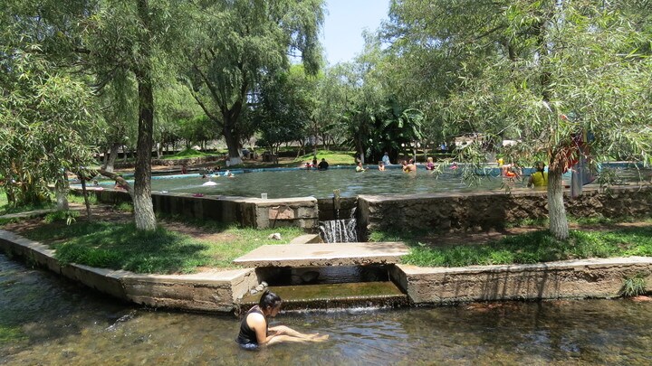 A scenic outdoor swimming area is surrounded by lush greenery and trees. The clear water reflects sunlight as people enjoy their time in the water. A small waterfall is visible, flowing gently into the pool, creating a serene ambiance.