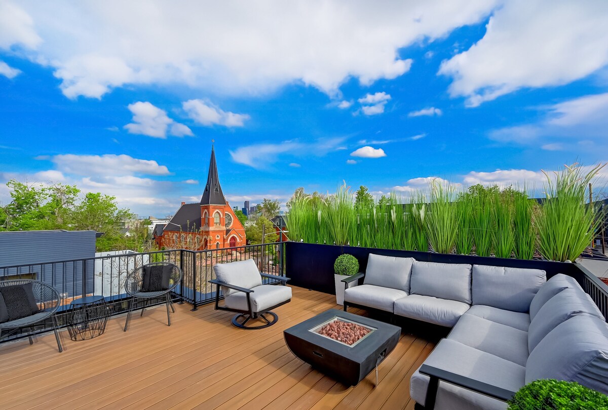 A rooftop deck features comfortable seating arranged around a central fire pit, offering stunning views of the skyline and surrounding greenery. The space is complemented by a backdrop of a historic church with a tall spire against a bright blue sky.
