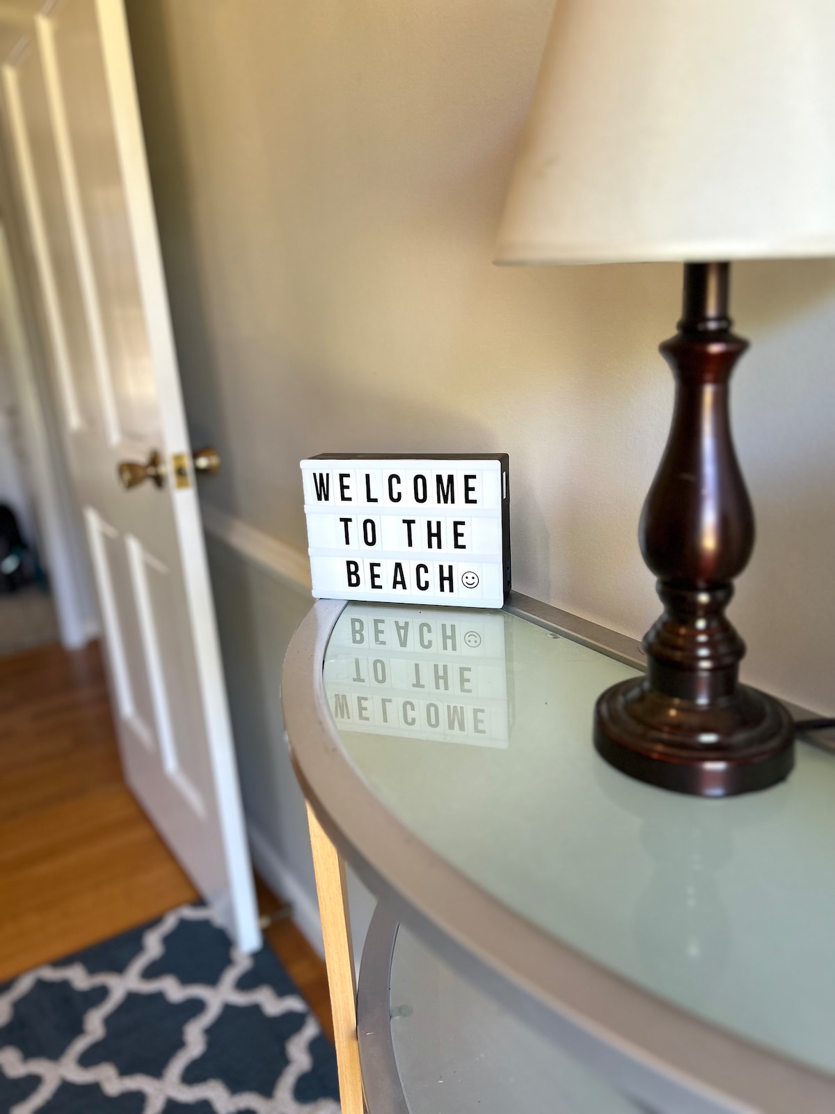 A decorative light box displays the message 'WELCOME TO THE BEACH' alongside a stylish lamp on a polished surface. The background features a softly hued wall and a partially visible wooden door, contributing to the inviting atmosphere of the space.