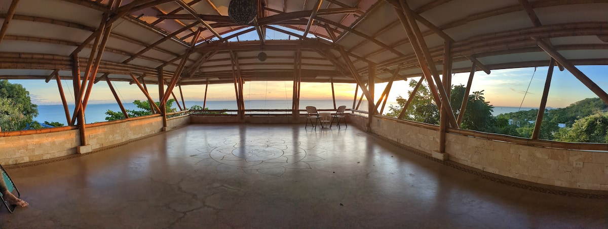 An expansive open-air bamboo deck features a polished concrete floor, offering unobstructed views of the ocean and sunset. Large windows frame the serene landscape, while a few chairs are positioned to take in the changing sky colors, providing a calm space for relaxation or activities.