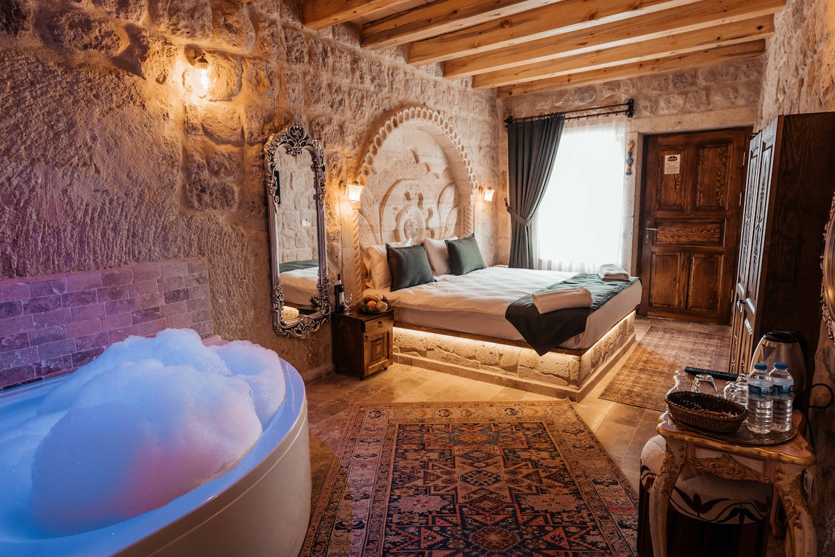 A charming room with natural stone walls features a luxurious jacuzzi, inviting relaxation. A spacious bed is placed next to a window dressed with dark curtains. An ornate mirror and a traditional area rug add character to the cozy atmosphere.
