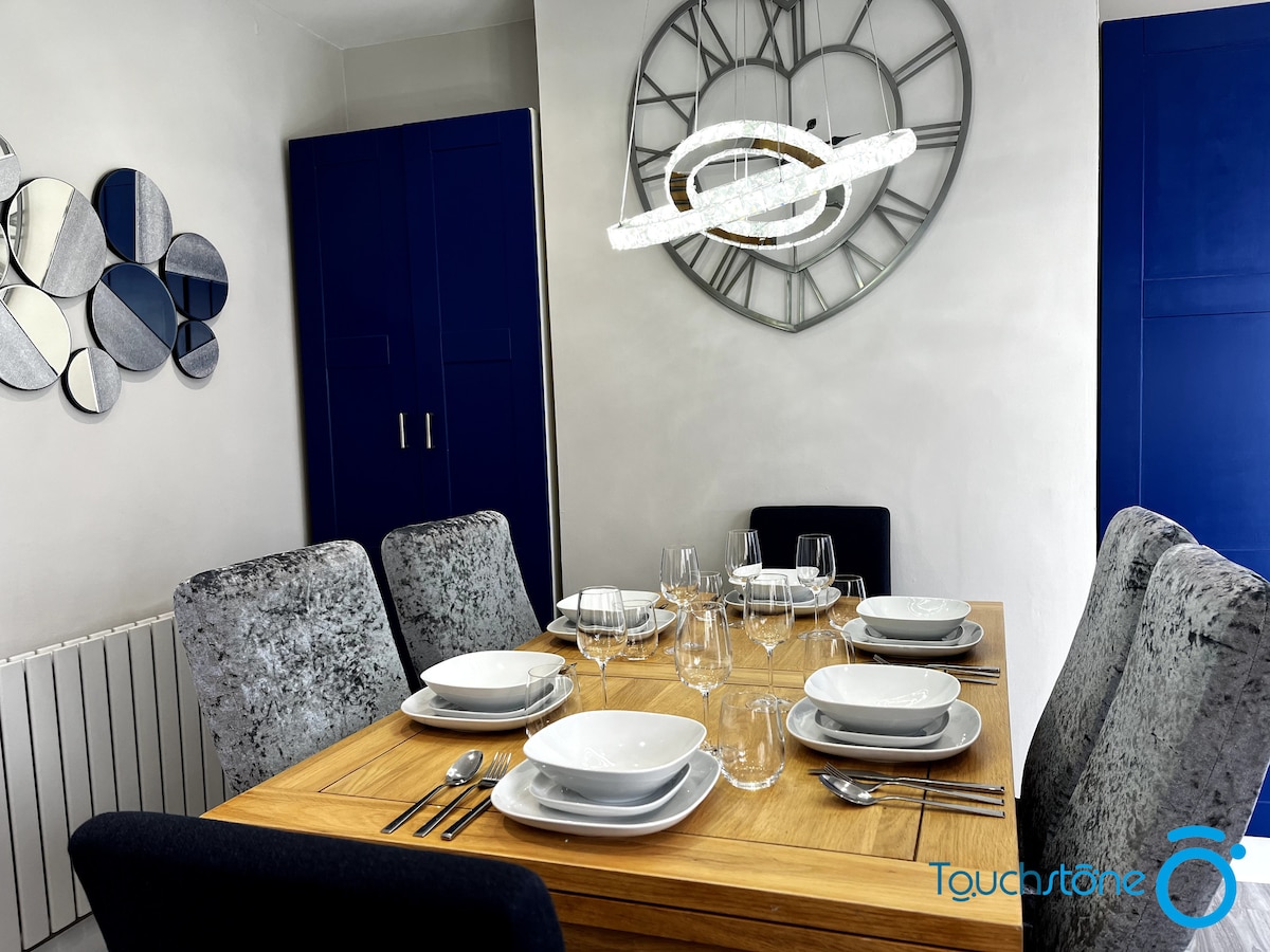 A dining area is shown, featuring a wooden table set with white plates, crystal glasses, and cutlery. Plush upholstered chairs surround the table, and a decorative wall clock complements the modern aesthetic. Wall-mounted mirrors reflect light and enhance the spacious feel of the room.