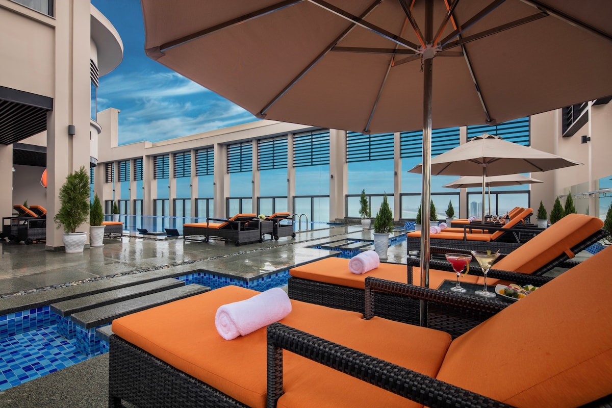 The rooftop pool area features comfortable lounge chairs with vibrant orange cushions, shaded by large umbrellas. A small pool reflects the sky, while potted plants add greenery. Refreshing drinks are seen on nearby tables, inviting relaxation and enjoyment of the panoramic views.