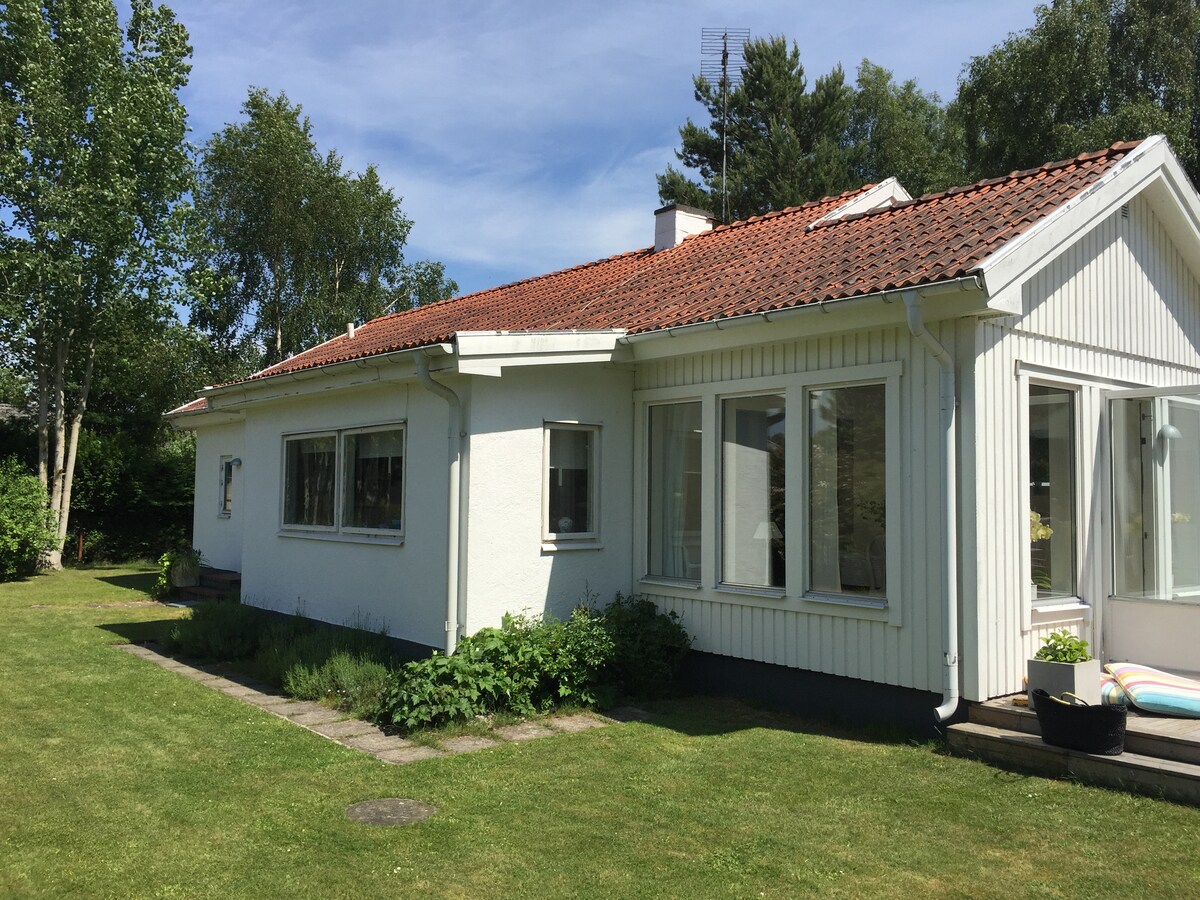 A single-story house is shown, featuring a white exterior with a red-tiled roof. Large windows allow natural light to enter the living spaces. A well-kept garden surrounds the house, with lush green grass and leafy plants enhancing the serene outdoor setting.
