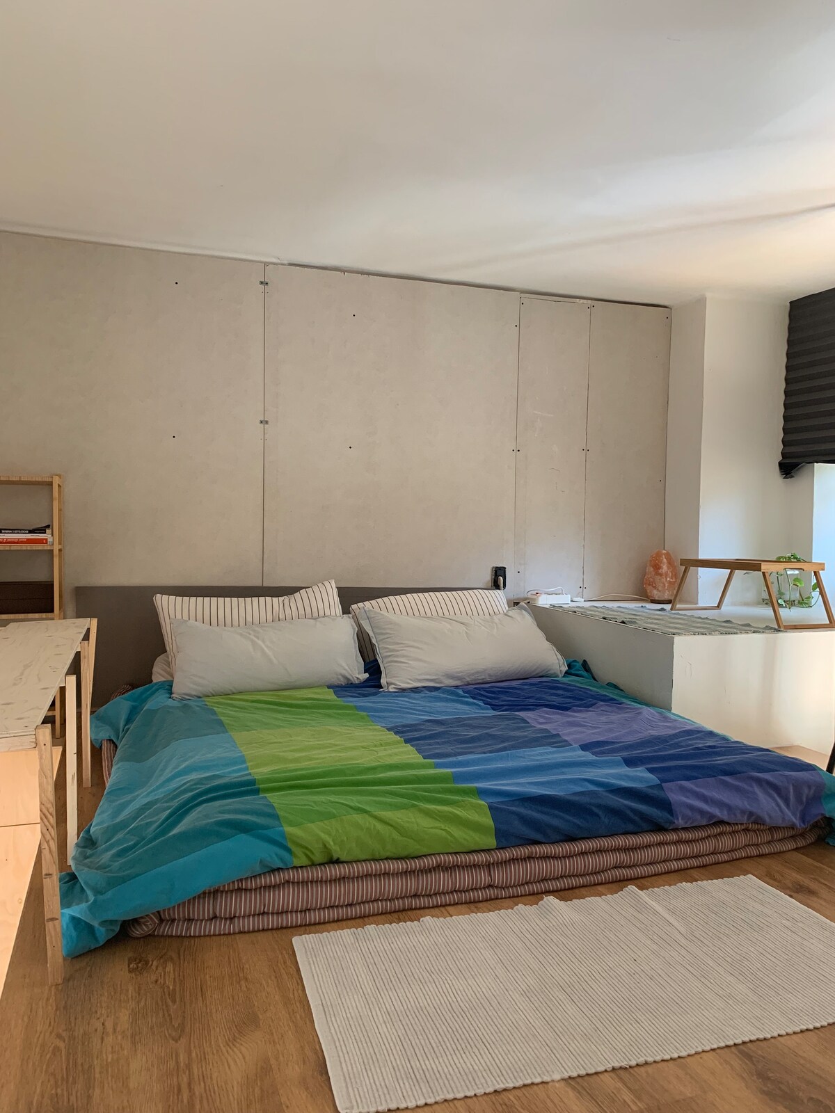 A spacious sleeping area features a large wool futon bed covered with a colorful blanket in shades of blue and green. A small area rug lies on the wooden floor, complementing the light walls and a simple side table. Natural light enters from a nearby window.