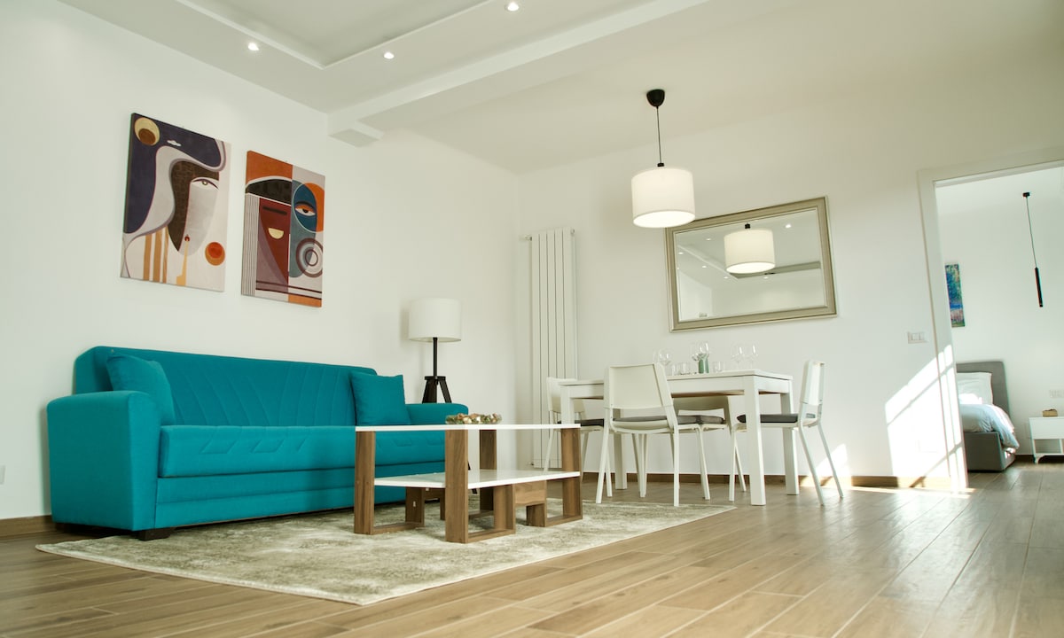 A spacious living area is highlighted by a bright blue sofa and an elegant coffee table, set upon a soft area rug. Light wooden floors complement a dining table with white chairs. Abstract art pieces add a touch of color to the white walls.
