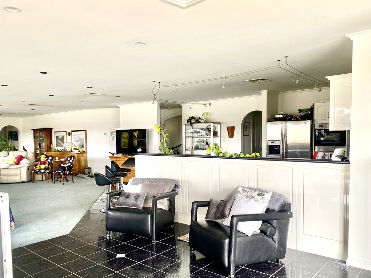 An open-plan living area is showcased, featuring a sleek kitchen with modern appliances and a spacious breakfast bar. Comfortable seating arrangements are included, with black chairs and light grey sofas positioned for relaxation. Large windows allow natural light to fill the space, enhancing the airy atmosphere.