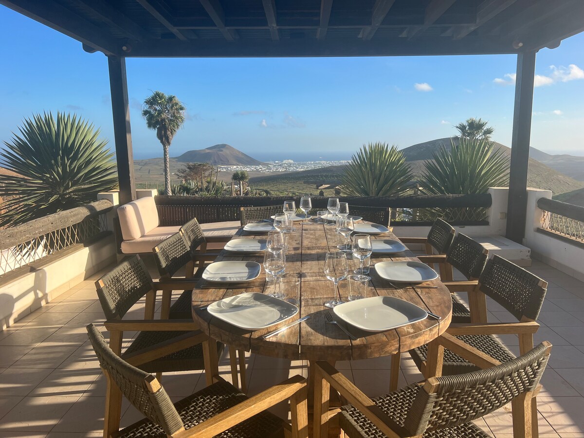 An outdoor dining area features a large wooden table surrounded by woven chairs, set with plates and glassware. Views of rolling hills and the coastline are visible in the background, complemented by palm trees and a clear sky.