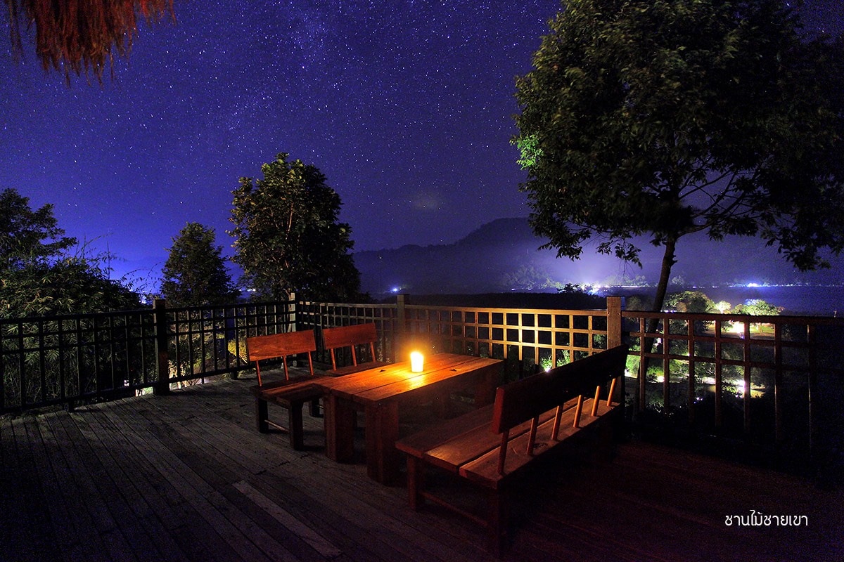 An outdoor seating area is illuminated by a single candle, inviting visitors to enjoy the starry night sky. Wooden benches and a table are positioned on a deck, surrounded by trees and distant mountains, creating a serene atmosphere.