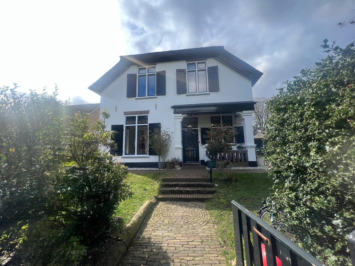 A charming two-story house is framed by lush greenery, featuring dark shutters and a welcoming entrance. A pathway made of cobblestones leads to the front door, creating a pleasant approach. The exterior is painted in light tones, complemented by well-maintained shrubs and trees.