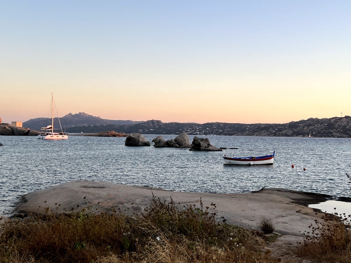 A tranquil coastal scene is captured at sunset, featuring a calm bay with several boats anchored. Large rocks dot the water's edge, while gentle waves lap against the shoreline. Soft hues of orange and pink blend in the sky, enhancing the serene atmosphere.
