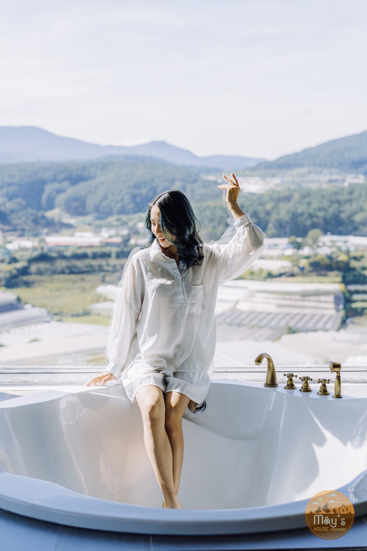 The image features a spacious bathtub positioned by a large window, offering stunning views of the surrounding mountains and valleys. Natural light fills the area, enhancing the serene ambiance. A person is seated on the edge of the tub, dressed in a light, comfortable outfit.