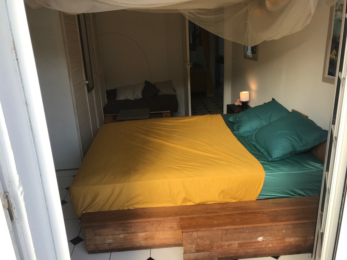 A cozy bedroom is displayed, featuring a spacious bed dressed in vibrant bedding with a yellow and teal color scheme. Light filters in from an open doorway, illuminating the simple wooden furniture and soft lighting from a bedside lamp. A corner seating area is also visible.