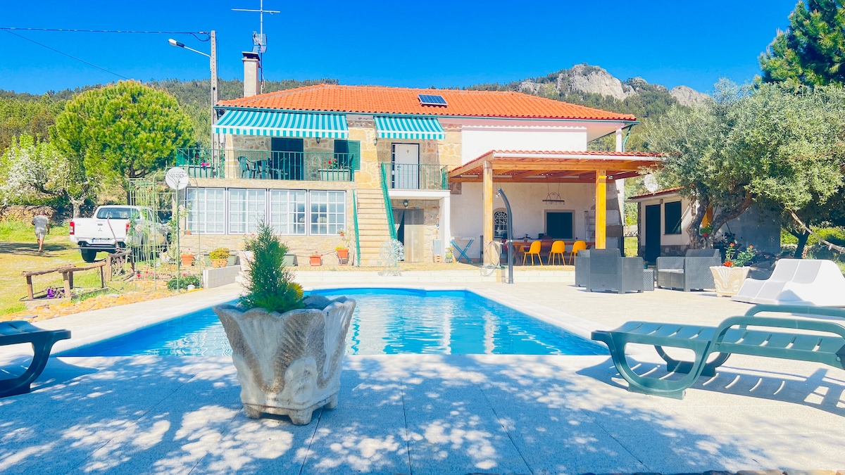 A stone house with a red-tiled roof is set against a clear blue sky, featuring a swimming pool at the forefront. A covered seating area is visible to the right, with several chairs arranged around a table, offering a serene outdoor space surrounded by greenery.