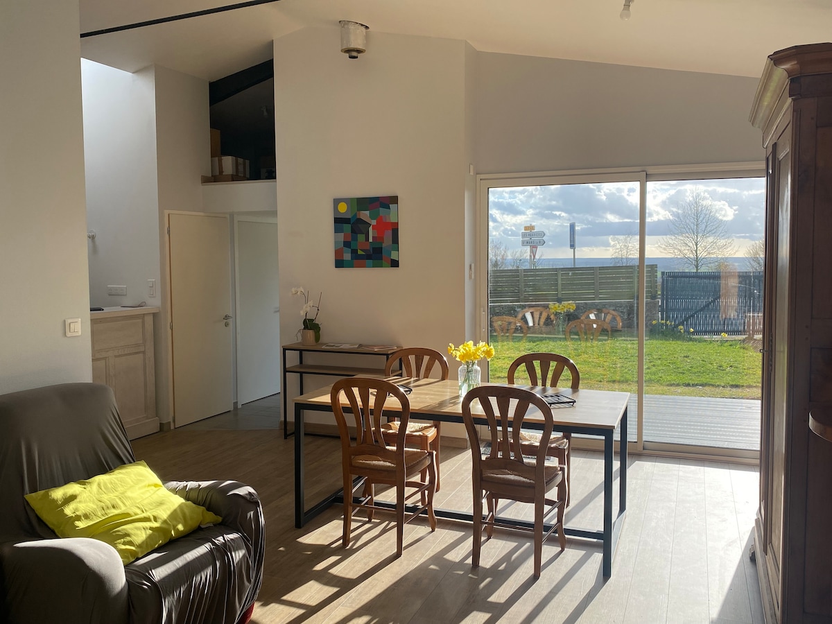 A bright dining area features a large table surrounded by four wooden chairs, with natural light streaming in through sliding glass doors. A comfortable armchair is positioned nearby, and colorful artwork is mounted on the wall. Greenery is visible through the outdoor space.