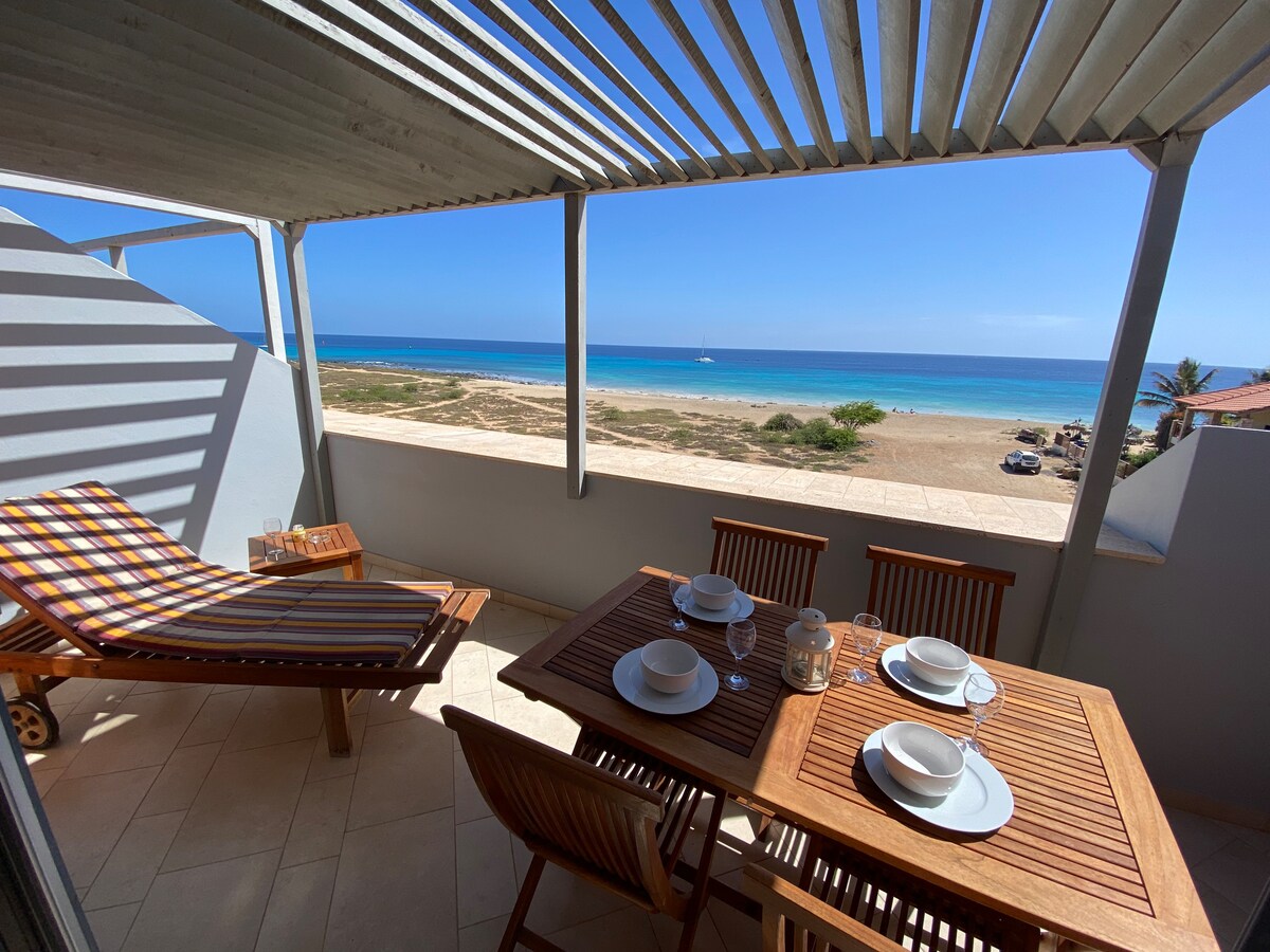 A shaded terrace is showcased, featuring a dining table set with plates and glassware. A striped lounge chair is positioned nearby, offering a relaxing space. The view encompasses the beach and sea, visible through the open space, with clear skies in the background.