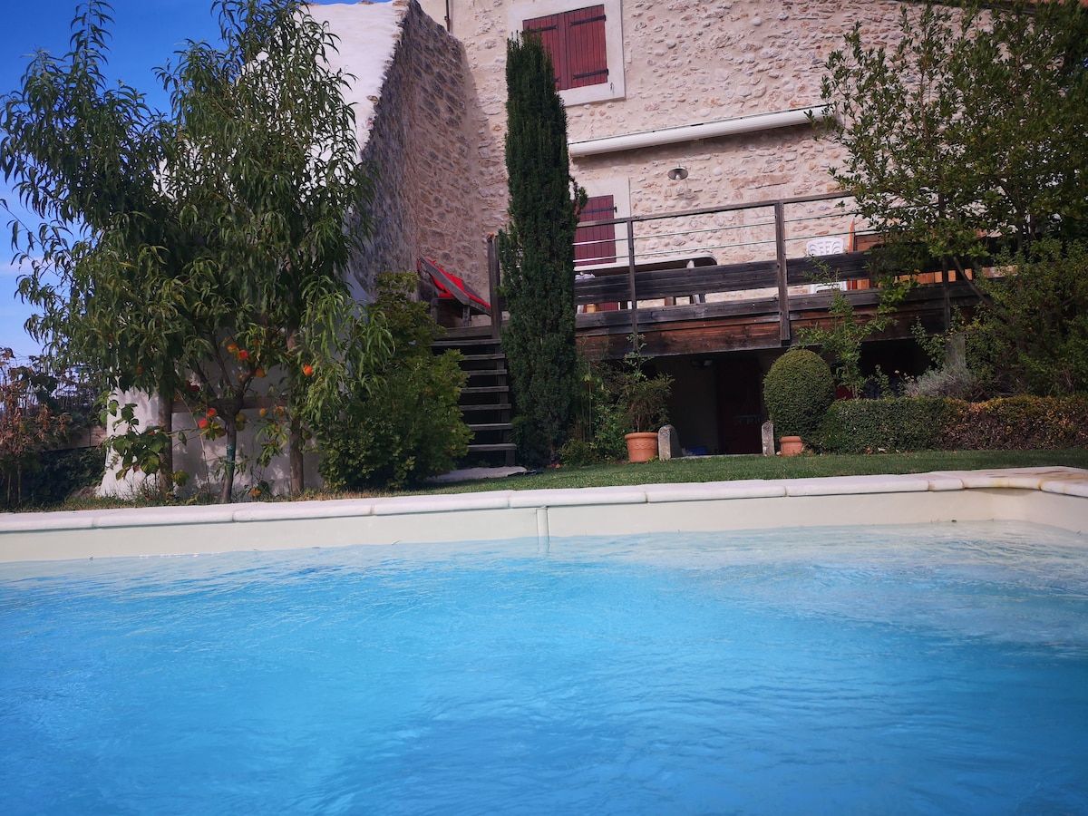 The image captures a shared swimming pool in the foreground, with clear blue water. A stone building with red window shutters is visible in the background, surrounded by greenery and potted plants. A wooden terrace is elevated, providing access to the building.